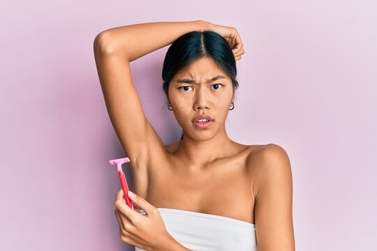 Young chinese woman wearing shower towel using razor clueless and confused expression. doubt concept.