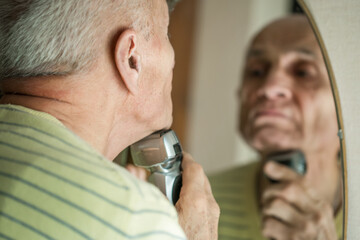 Gray haired senior man looking in mirror and shaving with electric razor