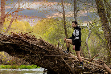 young man in sportswear and shorts climbing on a fallen tree trunk looking at the horizon with a stream and mountains in the background