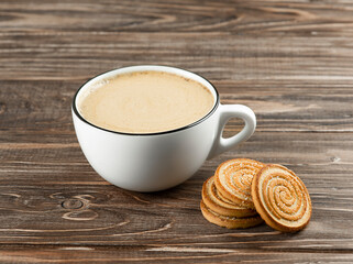 White cup with cappuccino coffee and cookies on a brown wooden background.