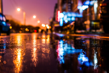 Rainy night in the big city, the street with bright shop windows. Close up view of a puddle on the asphalt level - obrazy, fototapety, plakaty