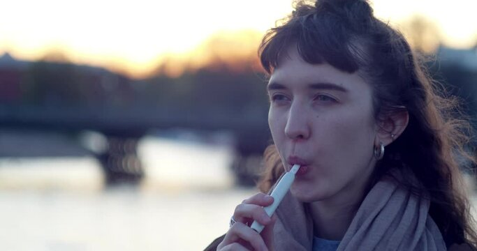 portrait of a girl smoking e-cigarettes in the street against the water and the sun leaving in the evening