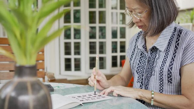 senior asian woman practicing writing chinese calligraphy at home patio