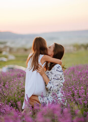 a woman and a baby girl with dark hair in white dresses are sincerely embracing in a lavender field . a landscape with hills and a pink sunset sky. mother, kid, sisters. children's day