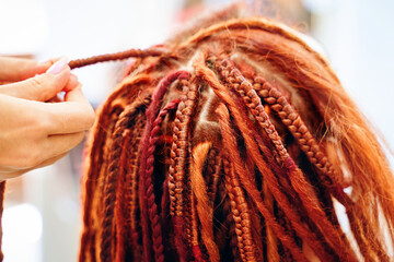 Close up of braiding process plait with colored kanekalon. Beauty salon services. Girl hairdresser weaves woman ginger dreadlocks. Stylish therapy professional care concept. Hippie style hairstyle.