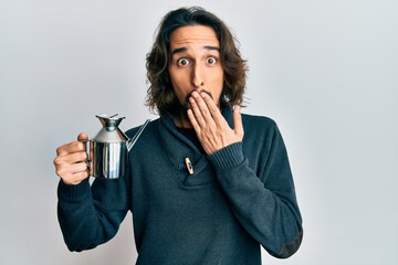 Young hispanic man holding olive oil can covering mouth with hand, shocked and afraid for mistake....