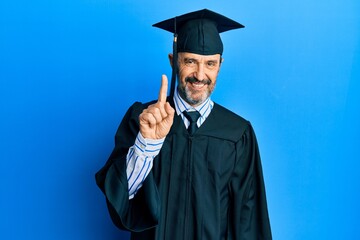 Middle age hispanic man wearing graduation cap and ceremony robe showing and pointing up with finger number one while smiling confident and happy.