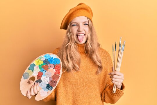 Beautiful young caucasian girl holding paintbrush and painter palette sticking tongue out happy with funny expression.