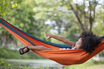 childhood and relax concept - little african american curly hair girl  lying down and enjoymenton on hammock color orange under big tree.