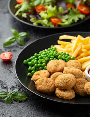 Fried crispy chicken nuggets with ketchup, french fries and green peas in black plate