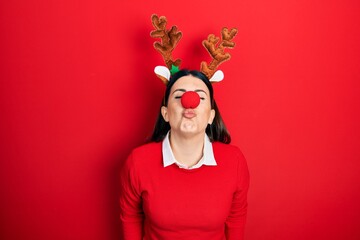 Young hispanic woman wearing deer christmas hat and red nose looking at the camera blowing a kiss on air being lovely and sexy. love expression.