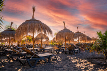 Chairs and umbrellas on the beach illuminated by sunrise against the colorful sky and clouds in Torviskas coast area in Tenerife island