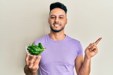 Young arab man holding bowl of green peppers smiling happy pointing with hand and finger to the side