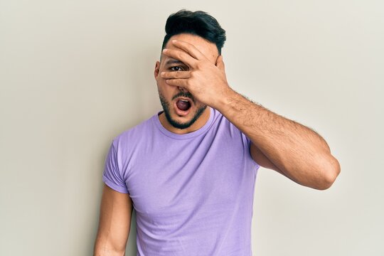 Young arab man wearing casual clothes peeking in shock covering face and eyes with hand, looking through fingers afraid