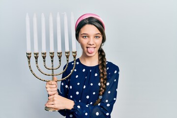 Young brunette girl holding menorah hanukkah jewish candle sticking tongue out happy with funny...