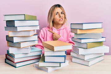 Young caucasian woman sitting on the table with books pointing aside worried and nervous with...