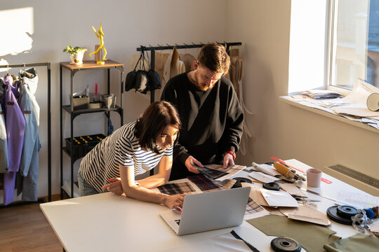 Team of fashion designers working together in studio. Young tailors man and woman choose fabric for suit collection, discuss new clothes material, creative process. Garment business, workshop interior