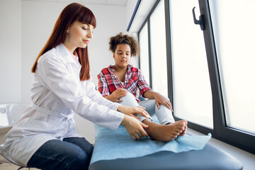 Trauma on the child's leg. Female Caucasian doctor orthopedist, examining foot of pretty mixed race...