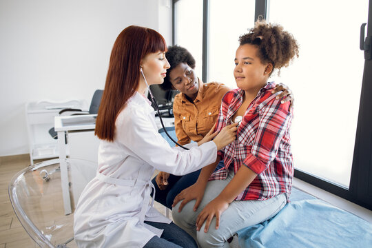 Flu, Cough, Asthma Concept. Female Pediatrician Doctor Listens To Lungs Of African-American 12-aged Girl With Stethoscope. Sick Teen Mixed Race Girl With Asthma, Supporting By Her Worried Mom