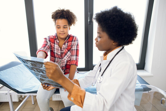 Young African woman doctor radiologist, shows x-ray tomography scan to cute child, pretty afro american curly schoolgirl, at modern light medical office