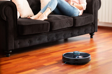 Faceless middle section of young woman relaxing on coach using automatic vacuum cleaner to clean the floor, controlling smart machine housework robot