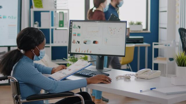 Black woman with disabilities and protection face masks working on computer in new normal workplace during pandemic. Multiethnic team in business financial office checking reports, analysing datas