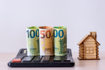 Rolled up paper money in denominations of 100 and 50 euros, on a calculator, against the background of a wooden house .The concept of the budget of the real estate market. Investment in construction.