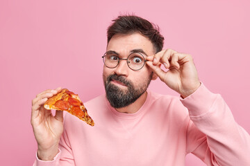Harmful delicious food. Handsome bearded man has tasty snack keeps hand on rim of spectacles holds...