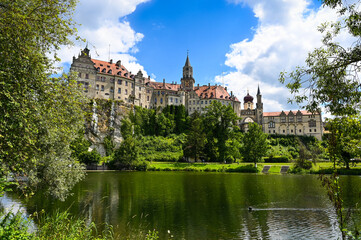 Scenic view of Sigmaringen Castle on a hill under a clear blue sky. The cityscape is reflecting in the water of the Danube river.