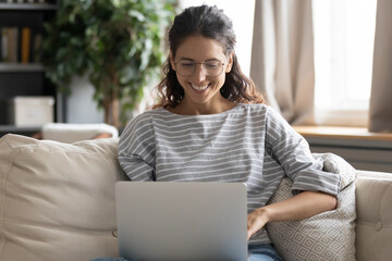Smiling millennial woman in glasses sit on sofa at home work online on modern computer. Happy young female look at laptop screen browse wireless internet or shop on web. Technology concept.