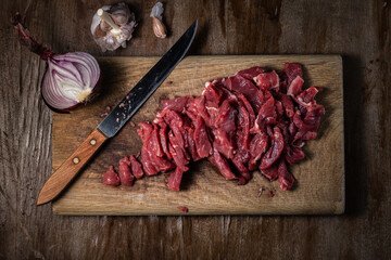 fresh raw meat cut into pieces in the process of cooking beef stroganoff lies on a wooden cutting board with a kitchen knife, onions and garlic on a background of old veneer