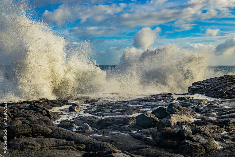 Wall mural Big waves on the rocky coast. White sea foam blown onto a beach. Sunny day on the ocean shore. Hawaii