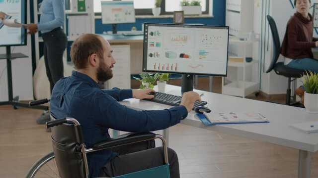 Paralized, immobilized, paralyzed business man writing financial report talking with colleague sitting at desk in wheelchair in modern startup business office Handicapped businessman working with team