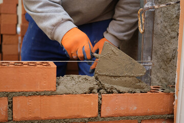 Construction worker putting cement on the brick wall. Man bricklaying. Mason laying bricks. - obrazy, fototapety, plakaty