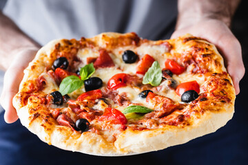 Male hands holding a pizza. Baked hot italian pizza with ham, black olives and tomatoes in hands