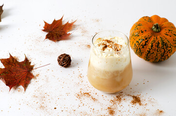Spicy pumpkin latte in a glass with cream and cinnamon. Concept of autumn warm drink with pumpkins and autumn leaves on white background with copy space.