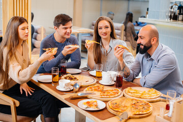 A group of young cheerful friends is sitting in a cafe talking and eating pizza. Lunch at the pizzeria.
