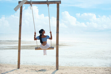 Young woman sitting on a swing over sea beach and blue sky. lifestyle outdoors in summer vacation travel trip.