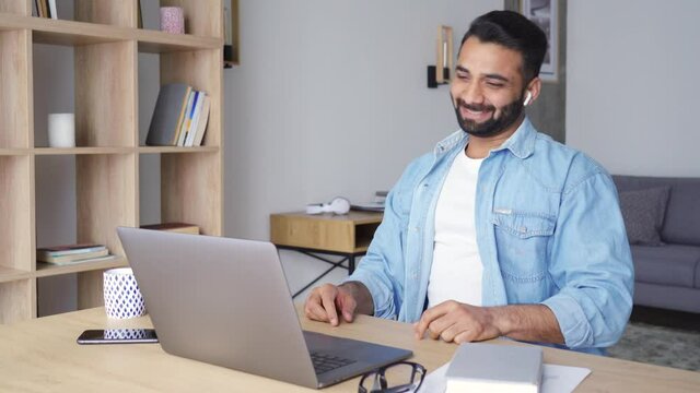 Young adult latin student guy leading online virtual training webinar using pc sitting at modern home office greeting, smiling and talking. Indian Hispanic man looking at computer screen on desk.