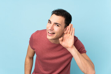 Young caucasian handsome man isolated on blue background listening to something by putting hand on the ear