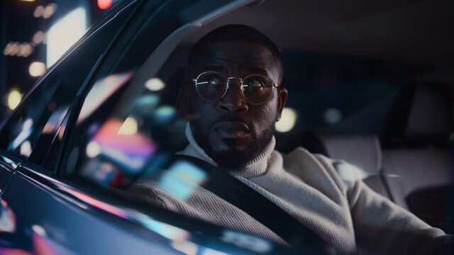 Stylish Black Man in Glasses is Commuting Home in a Backseat of a Taxi at Night. Handsome Male Passenger Looking Out of Window while in a Car in Urban City Street with Working Neon Signs.