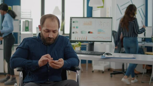 Handicapped, paralyzed immobilized invalid young employee browsing on smartphone during worktime typing, searching sitting in wheelchair. Diverse team discussing financial project, checking statistics