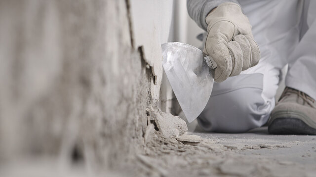 Construction worker hands with gloves working with spatula scrape off the plaster from the wall for house renovation, close up with rubble on floor