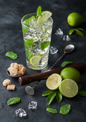 Glass of Mojito cocktail with ice cubes mint and lime on black board with spoon and muddler and fresh limes and cane sugar. Sparkling refreshing water.