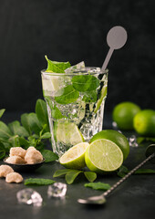 Glass of Mojito cocktail with ice cubes and disk stirrer,mint and lime on black board with spoon and fresh limes with cane sugar.
