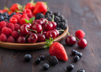 Fresh organic summer berries mix in round wooden tray on dark wooden table background. Raspberries, strawberries, blueberries, blackberries and cherries. Macro. Close up