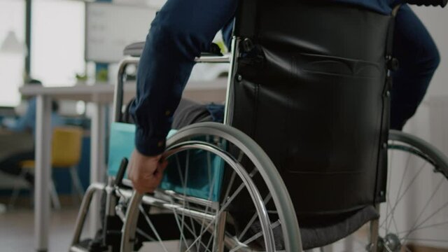 Close up independence handicapped man in wheelchair entering in company office, moving alone starting to work at financial project using professional computer after greeting colleagues