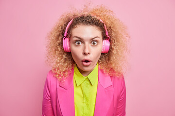 Surprised astonished woman with curly bushy hair looks amazed at camera listens music from playlist via wireless headphones dressed in bright formal outfit isolated over vivid pink background