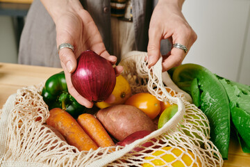 Hand of female holding fresh onion over bag with other vegetables