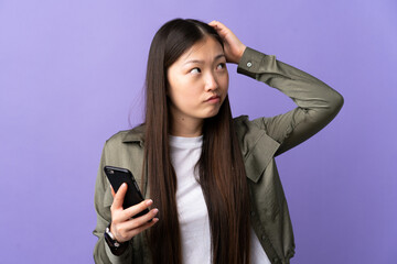 Young Chinese girl using mobile phone over isolated purple background having doubts and with confuse face expression